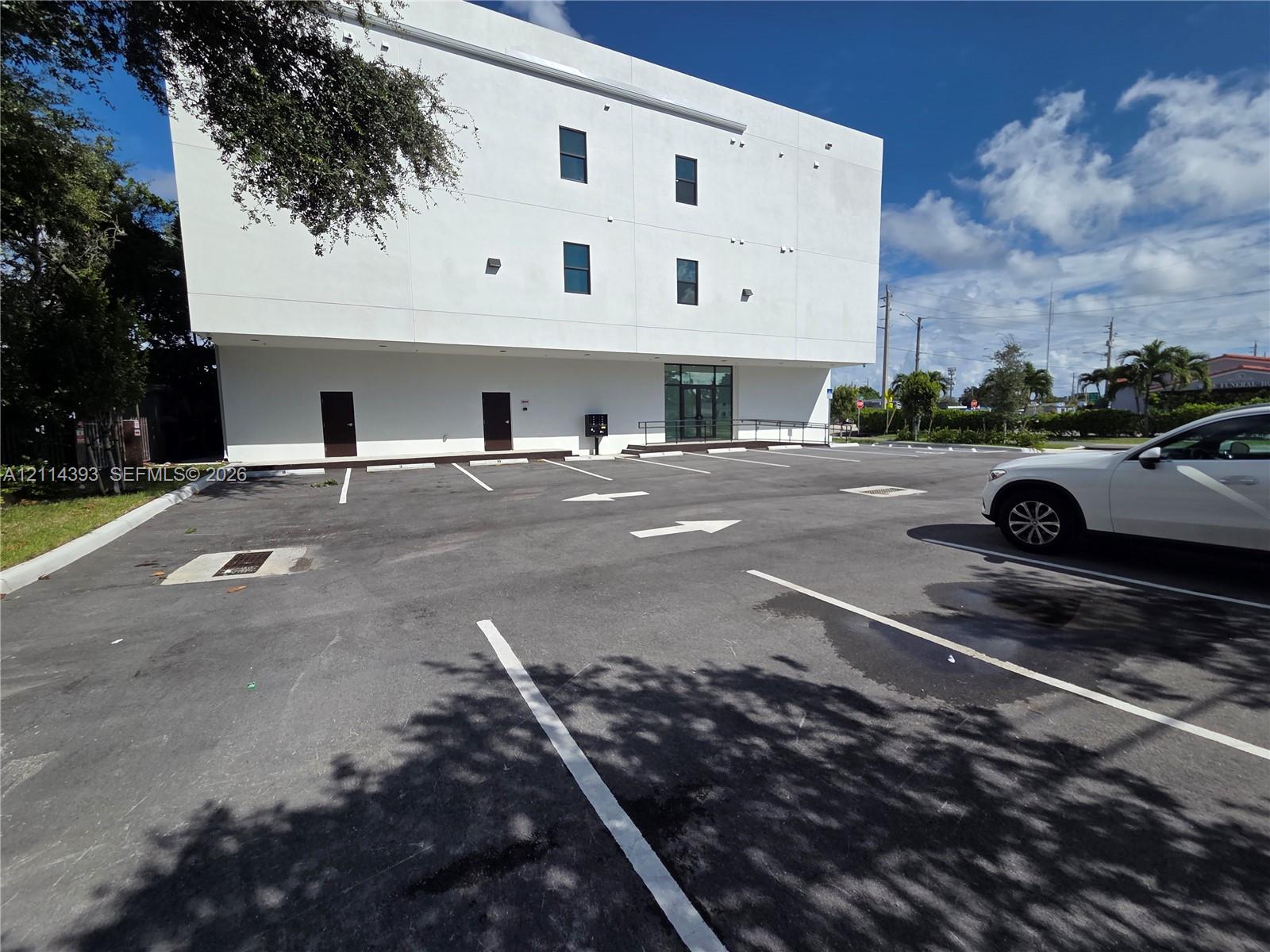 Three-story white stucco exterior featuring a paved asphalt parking lot with designated spaces, dual entry doors, and a glass-paneled entrance with an integrated ramp