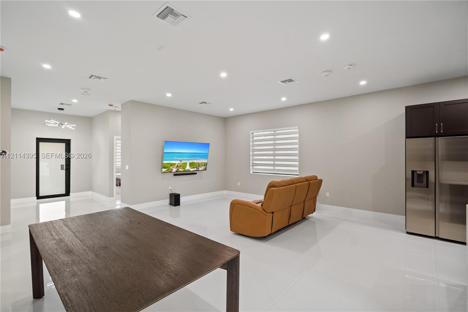 Open-concept interior featuring large-format tile flooring, recessed ceiling lighting, roller shades, and stainless-steel refrigerator with integrated cabinetry Open-concept interior featuring large-format tile flooring, recessed ceiling lighting, roller shades, and stainless-steel refrigerator with integrated cabinetry