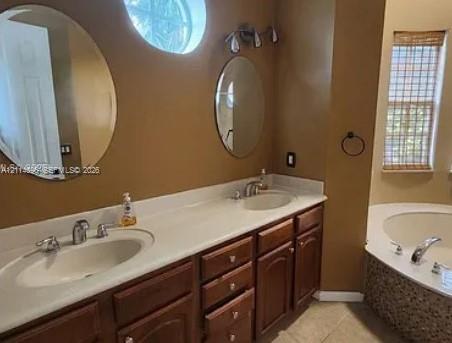 Dual-sink vanity with light-toned countertop, wooden cabinetry, oval wall-mounted mirrors, circular accent window, and jetted soaking tub featuring decorative tile surround