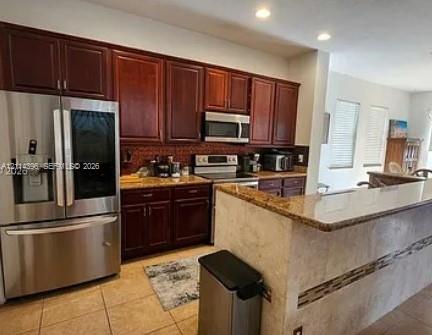 Kitchen featuring dark-toned cabinetry, stainless steel appliances, stone-finish countertops, a breakfast bar with a tile facade, and recessed ceiling lighting