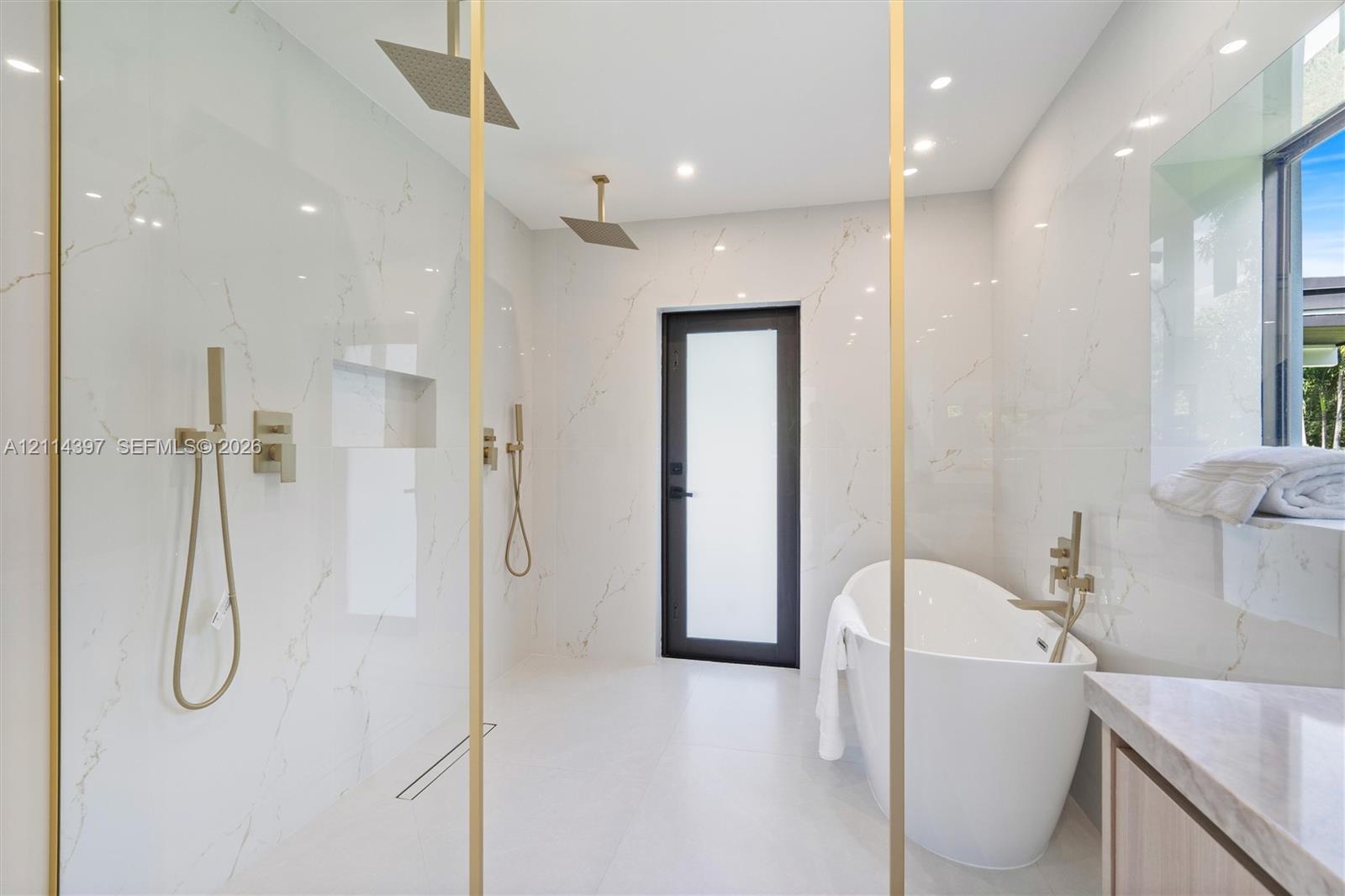 Walk-in wet room featuring oversized wall-to-wall stone-finish tile, dual ceiling-mounted rainfall showerheads, brass-finish fixtures, a freestanding soaking tub, and a built-in shower niche