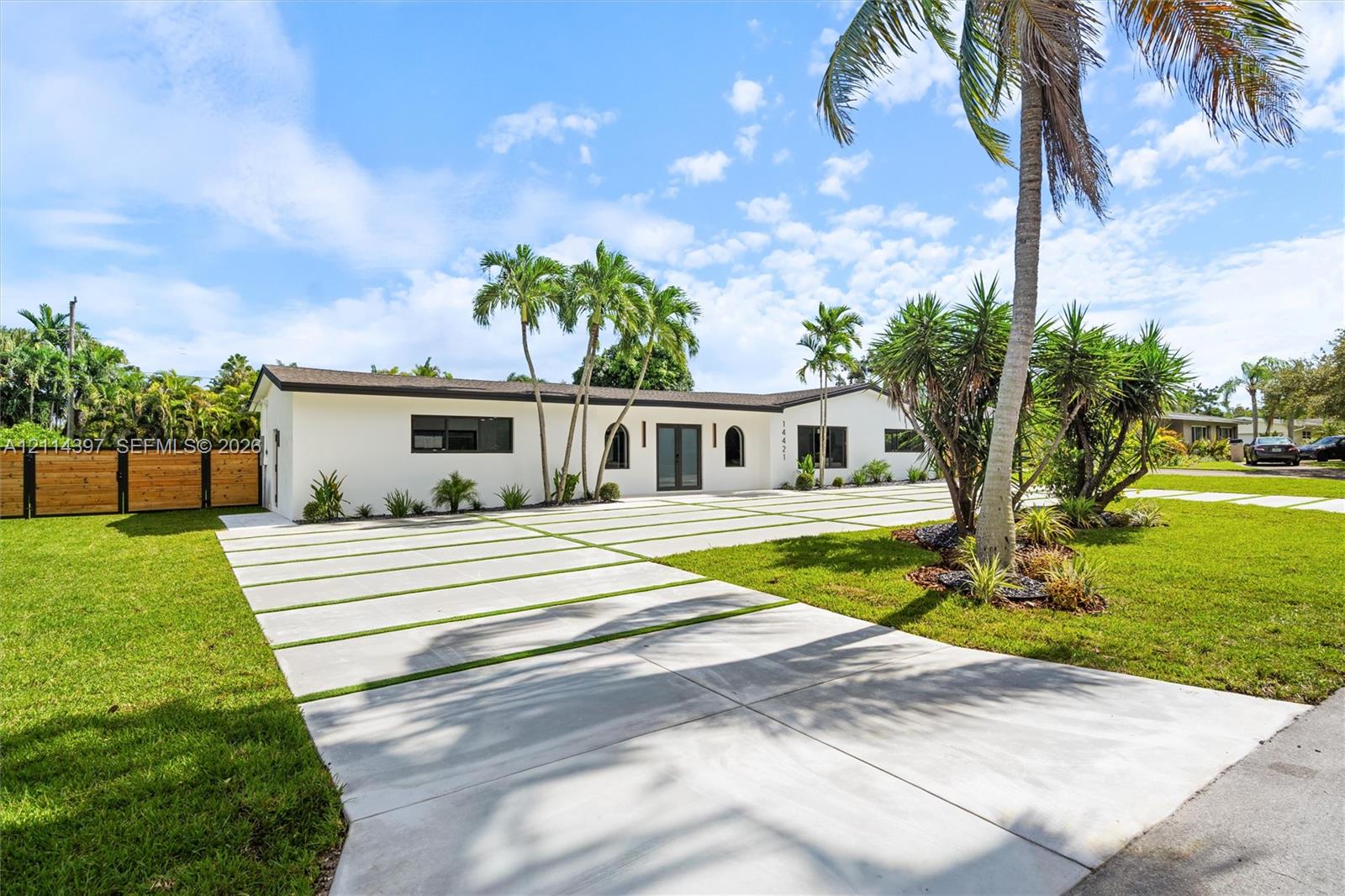 Single-story residence featuring a smooth stucco exterior, expansive concrete driveway with grass inlays, arched entryway, horizontal wood-slat privacy fencing, and mature palm landscaping