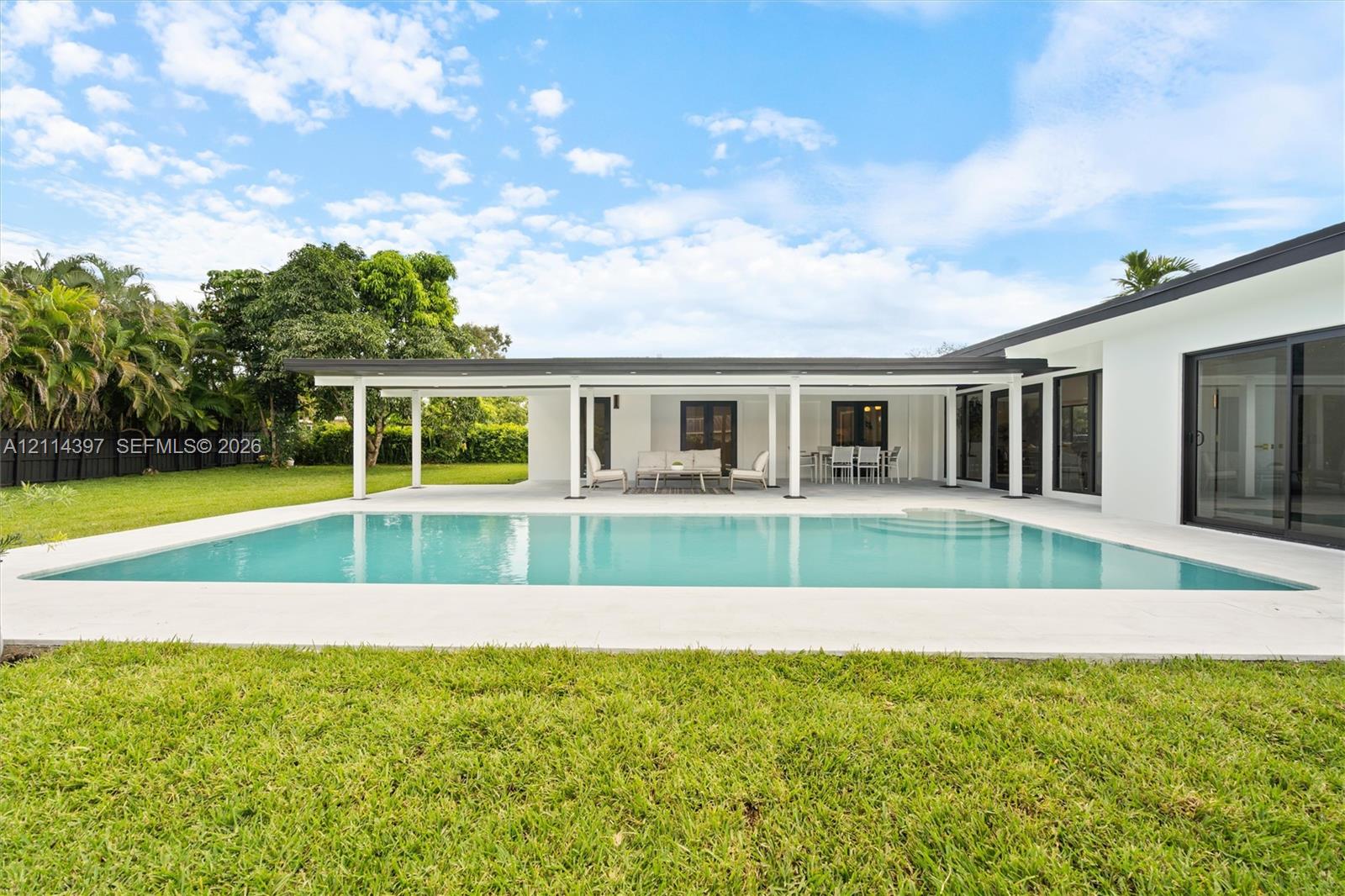 Backyard featuring a rectangular swimming pool, expansive concrete patio, covered lanai with structural pillars, sliding glass door panels, and manicured lawn