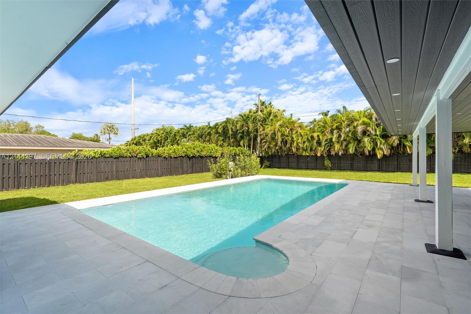 Rectangular in-ground pool featuring a tiled surround, covered patio area, and privacy fencing enclosing a lawn