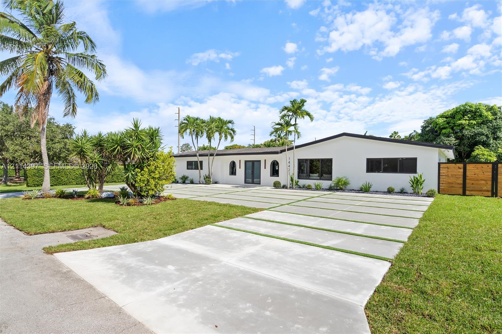 Single-story residence featuring a smooth white exterior, expansive concrete driveway with inset grass turf strips, mature palm trees, and horizontal wood-panel fencing