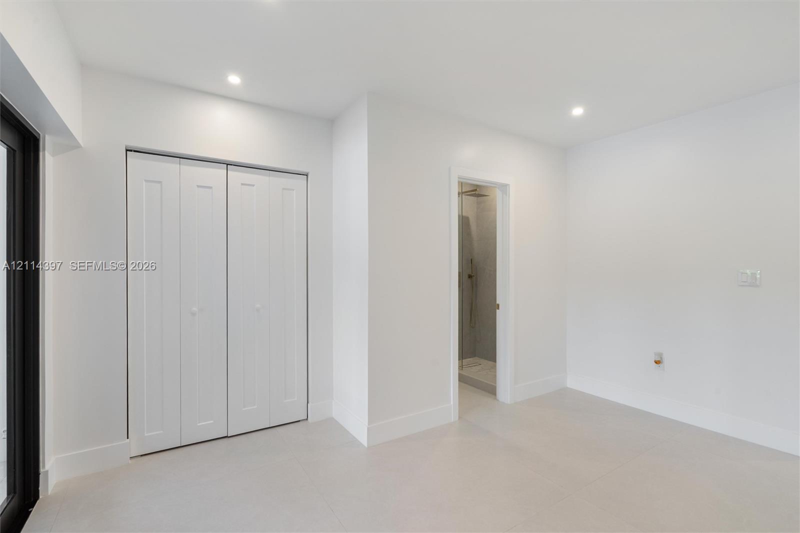 Interior space featuring large-format neutral floor tile, recessed ceiling lighting, a bi-fold closet, and a doorway revealing a tiled shower enclosure