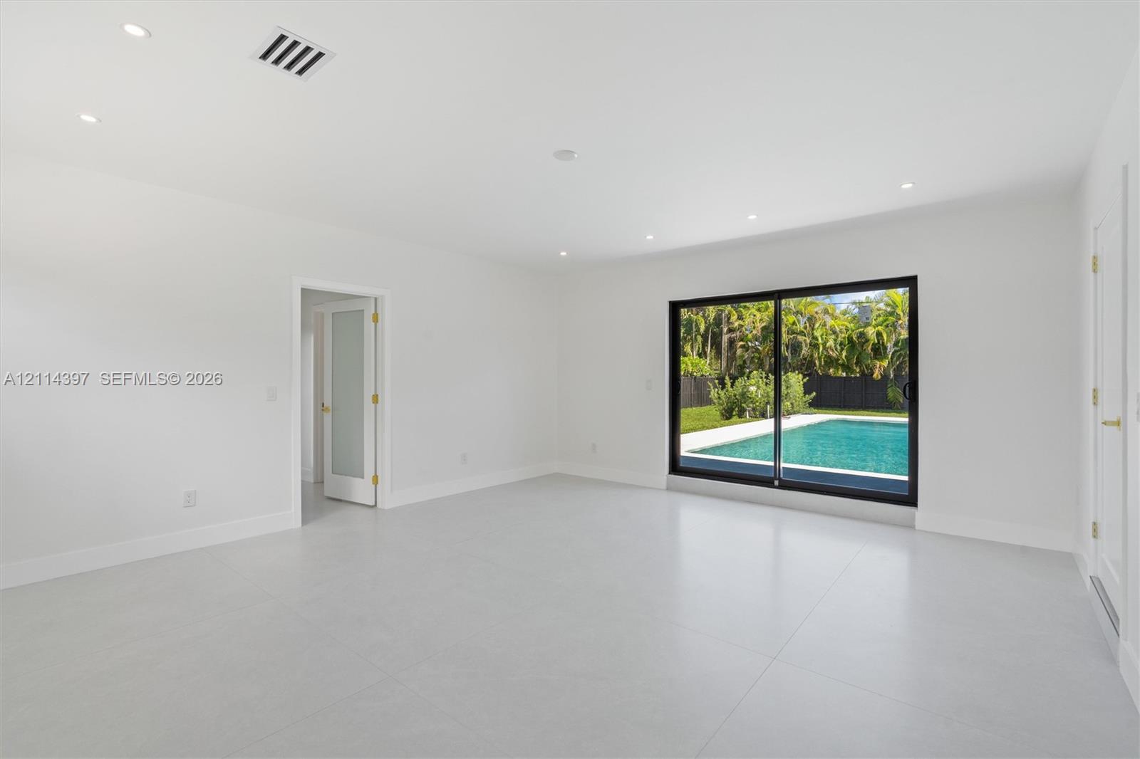 Open interior space featuring light-toned tile flooring, recessed ceiling lighting, a sliding glass door with a dark frame, and a frosted glass panel door