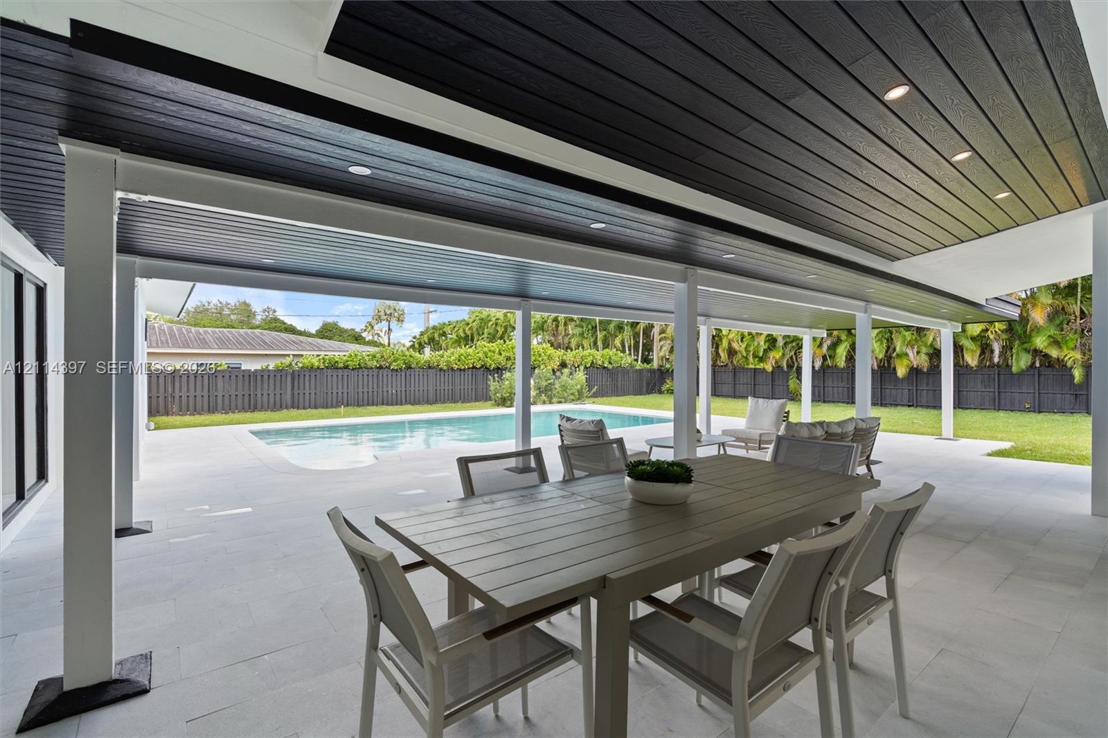 Covered outdoor patio featuring a dark-stained wood tongue-and-groove ceiling, recessed lighting, white support pillars, light-toned paved flooring, and an in-ground swimming pool