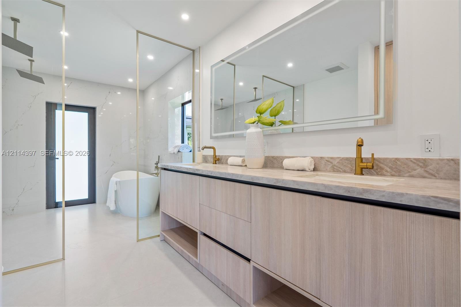 Dual-sink vanity with wood-finish cabinetry, natural stone countertop, brass fixtures, integrated LED-backlit mirror, and glass-enclosed wet room featuring a soaking tub