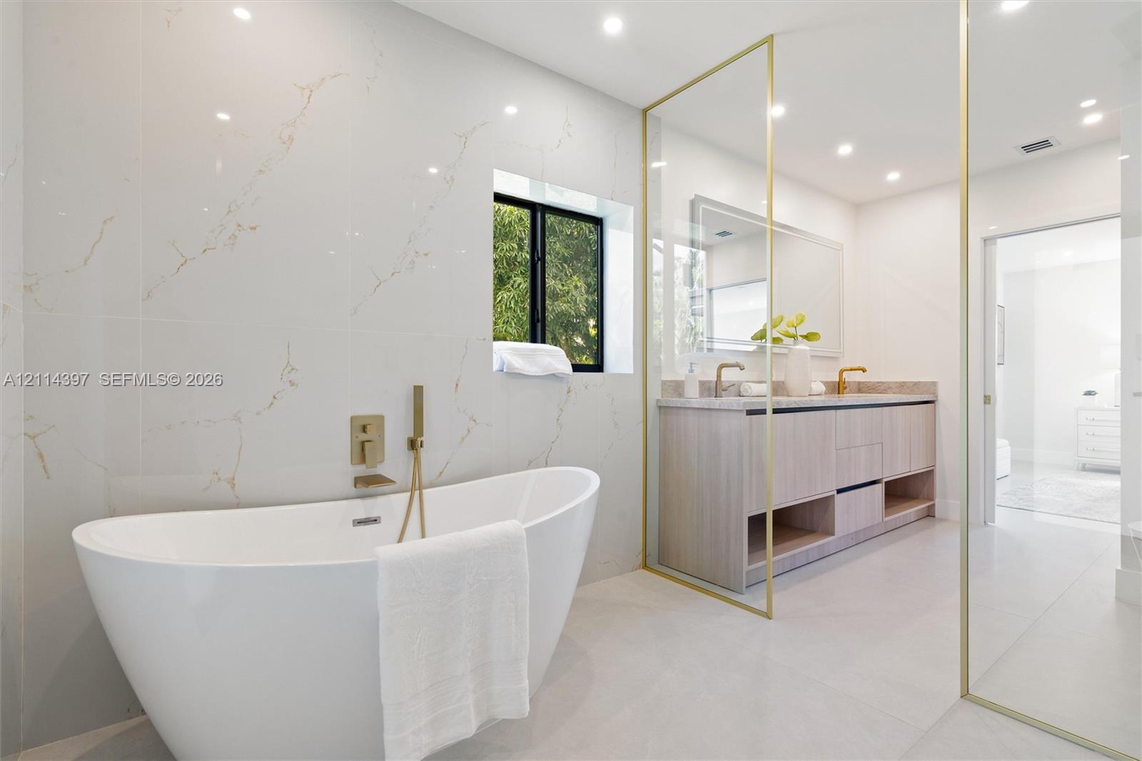 Freestanding soaking tub with gold-tone wall-mounted faucet, large-format tile wall cladding, wood-finish floating vanity with dual sinks, gold-trimmed glass enclosure, and recessed lighting