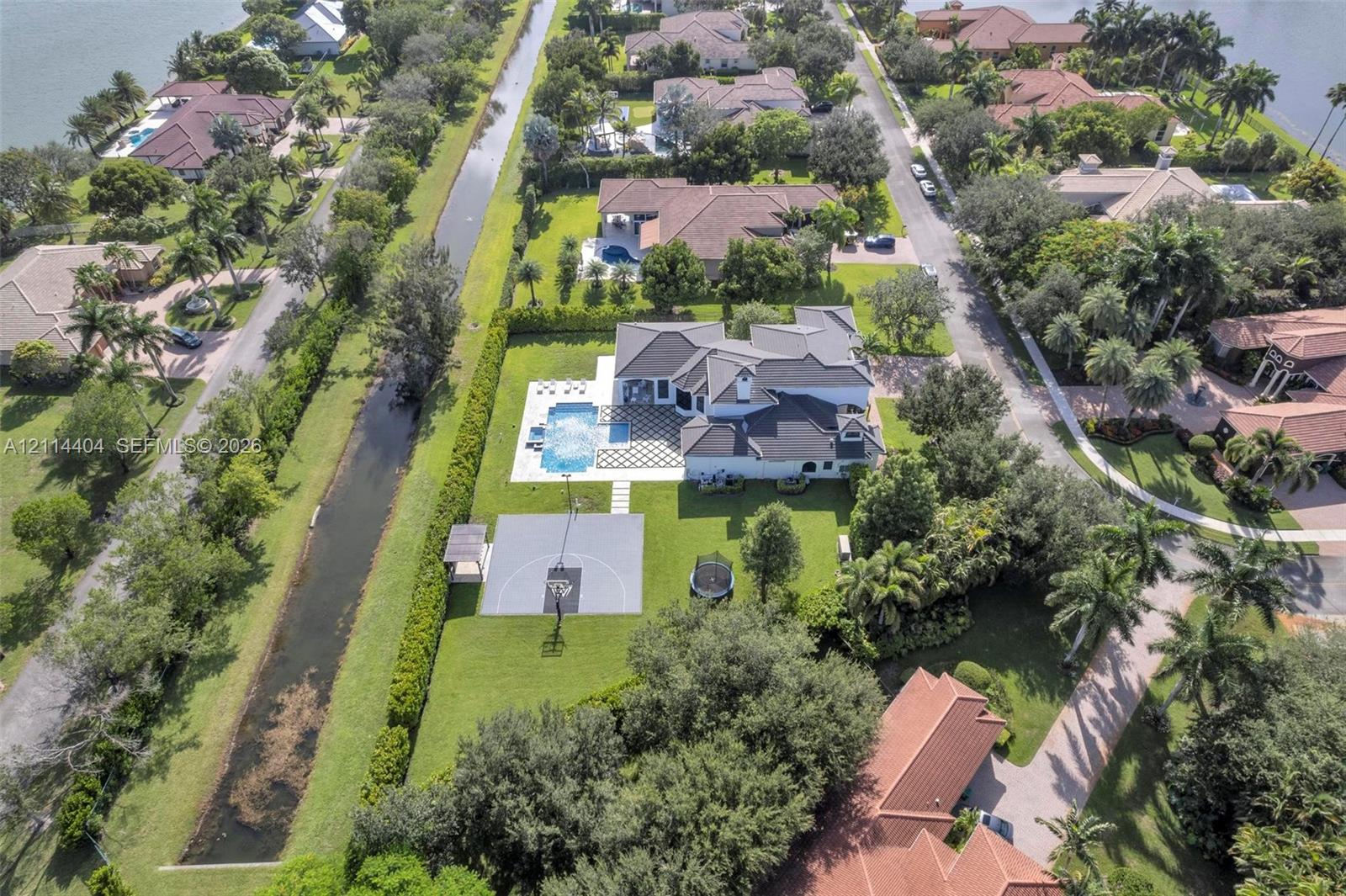 Aerial view of waterfront property featuring a swimming pool, paved patio, multi-use hardscaped court, and manicured landscaping