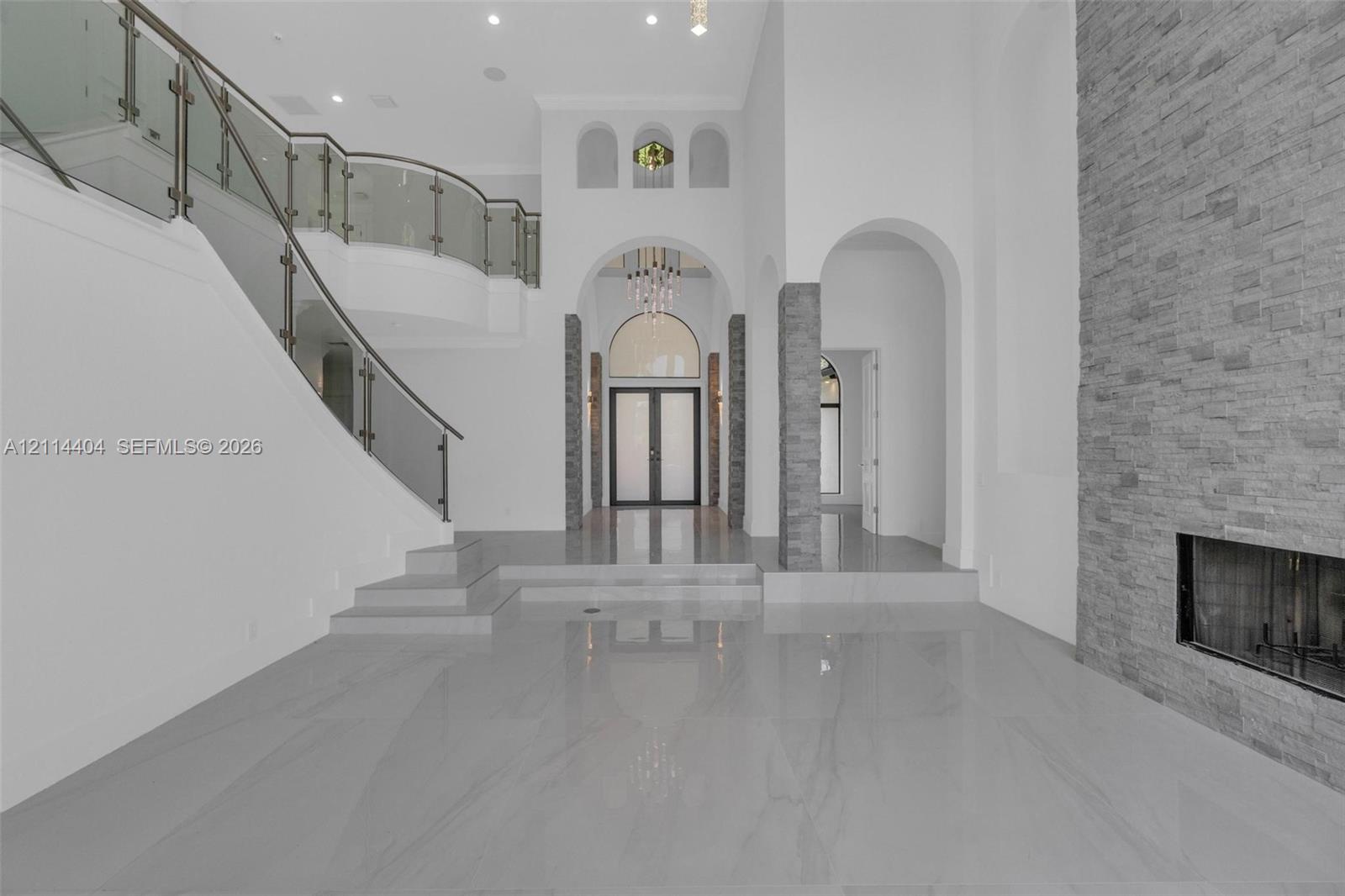 Grand foyer featuring polished stone flooring, a sweeping staircase with glass railings, soaring arched entryways, and a floor-to-ceiling stacked stone fireplace Grand foyer featuring polished stone flooring, a sweeping staircase with glass railings, soaring arched entryways, and a floor-to-ceiling stacked stone fireplace