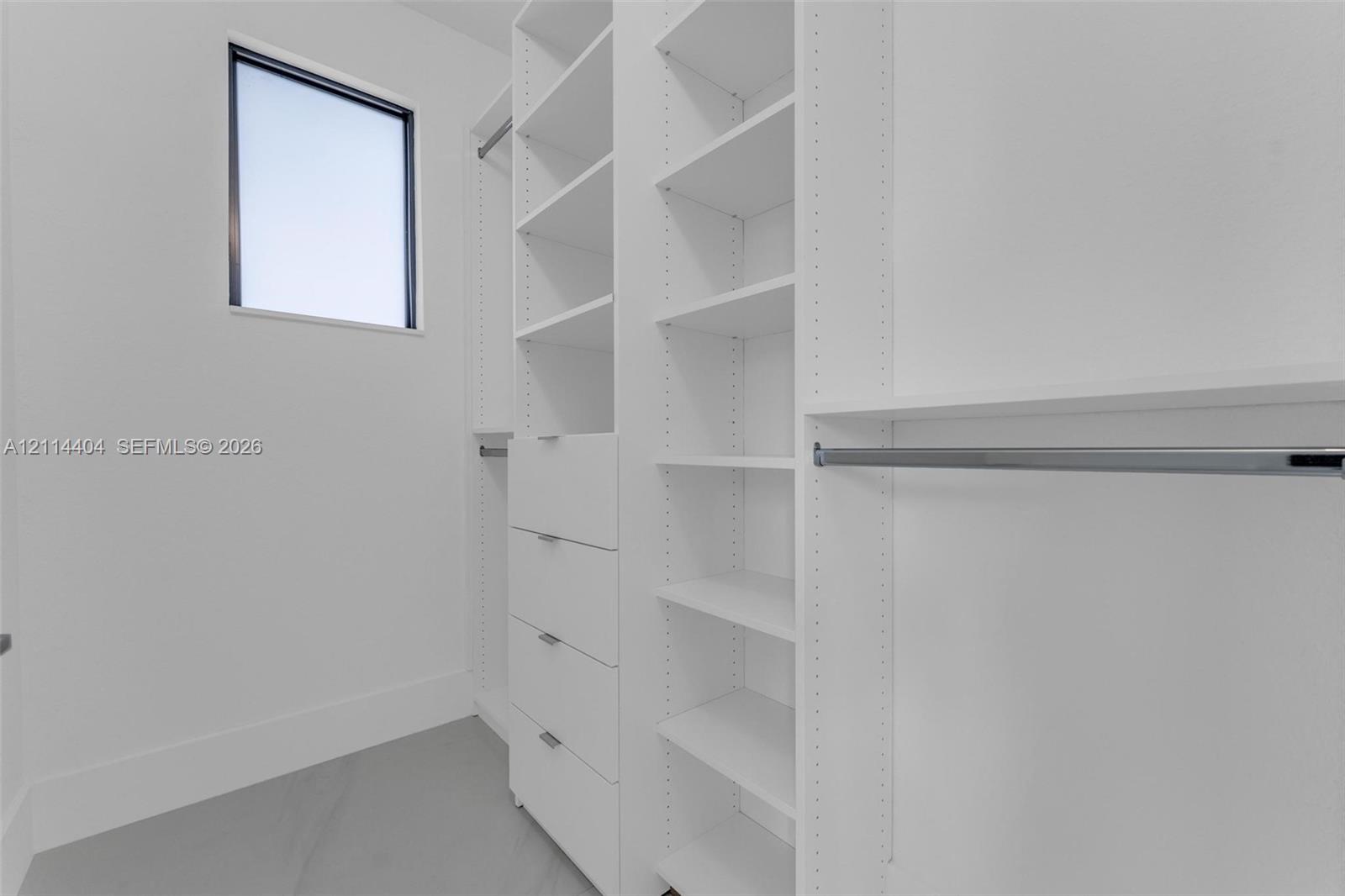 Custom walk-in closet featuring white built-in shelving, a four-drawer dresser unit, metallic clothing rods, and a rectangular window with a dark frame