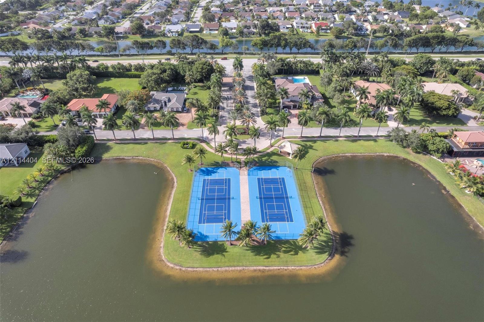 Dual tennis courts situated on a peninsula surrounded by water, featuring perimeter fencing and landscaped palm trees