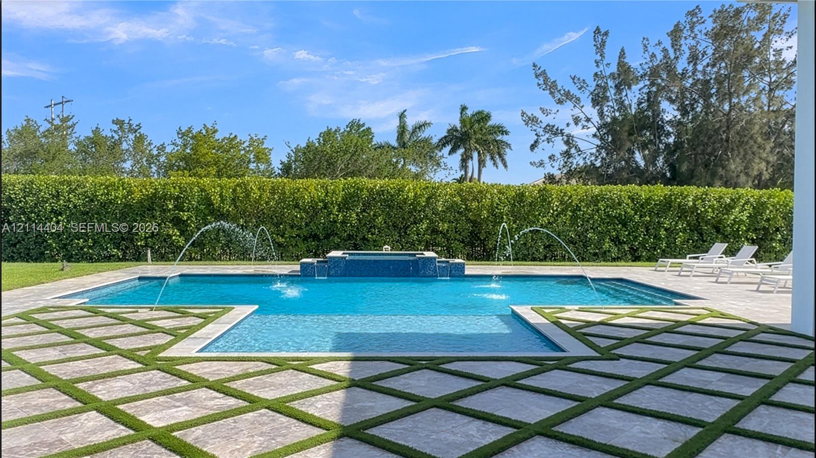Swimming pool with built-in water features and integrated spa, surrounded by stone paver decking with grid-pattern turf inlay and mature perimeter hedging