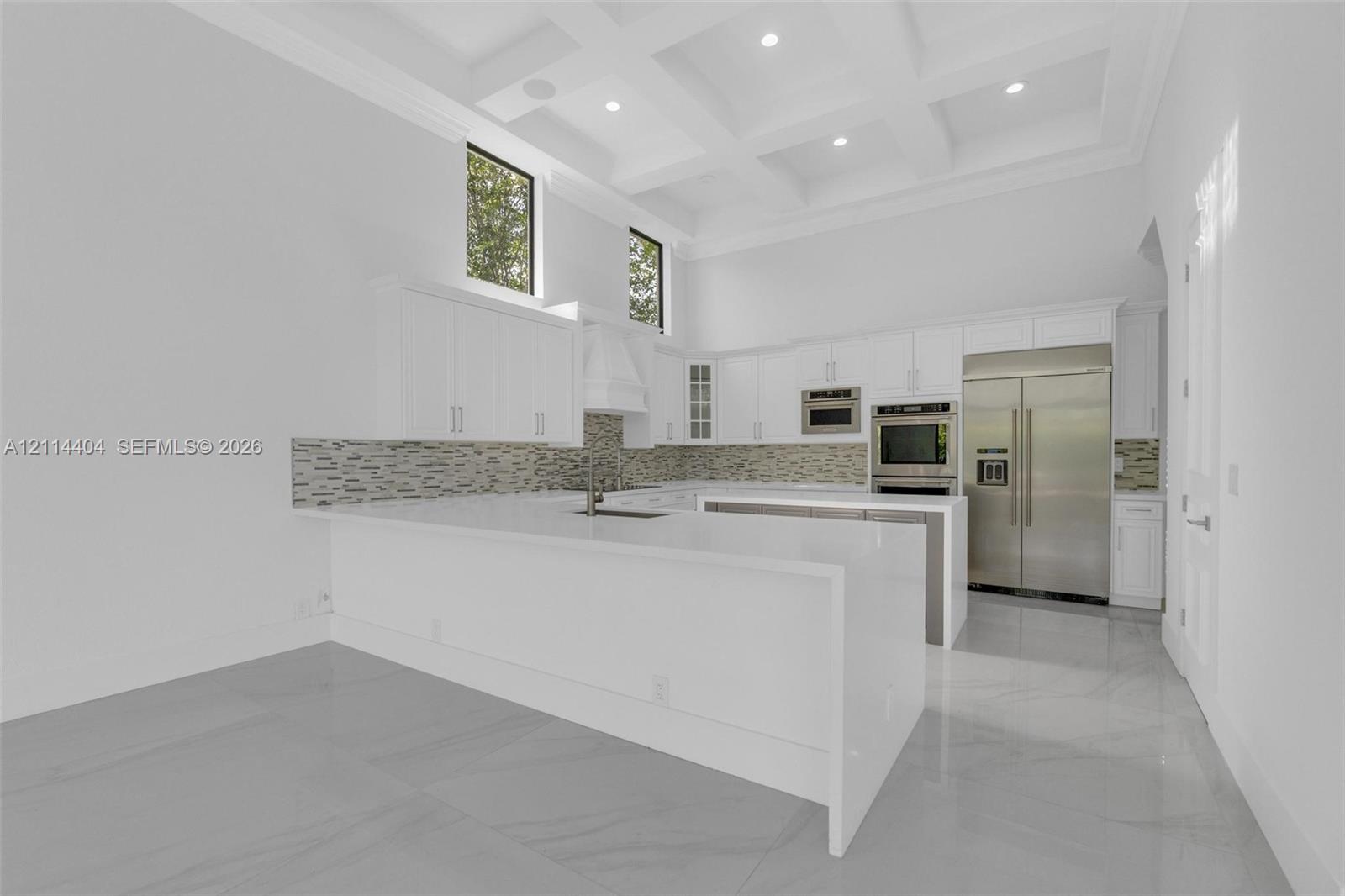 Kitchen featuring a coffered ceiling, waterfall-edge quartz island, stainless steel appliances, stacked-stone backsplash, and cabinetry with integrated recessed lighting
