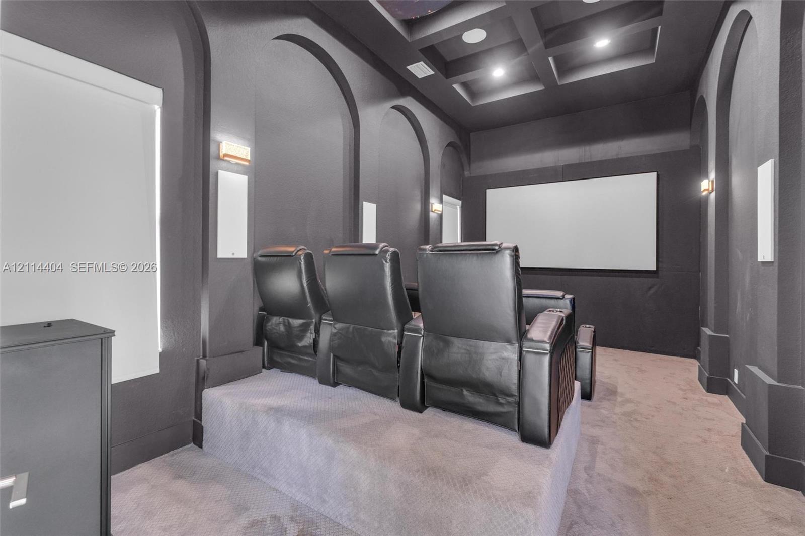 Media room featuring a coffered ceiling, tiered flooring, arched wall alcoves, integrated wall-mounted lighting, and a fixed projection screen