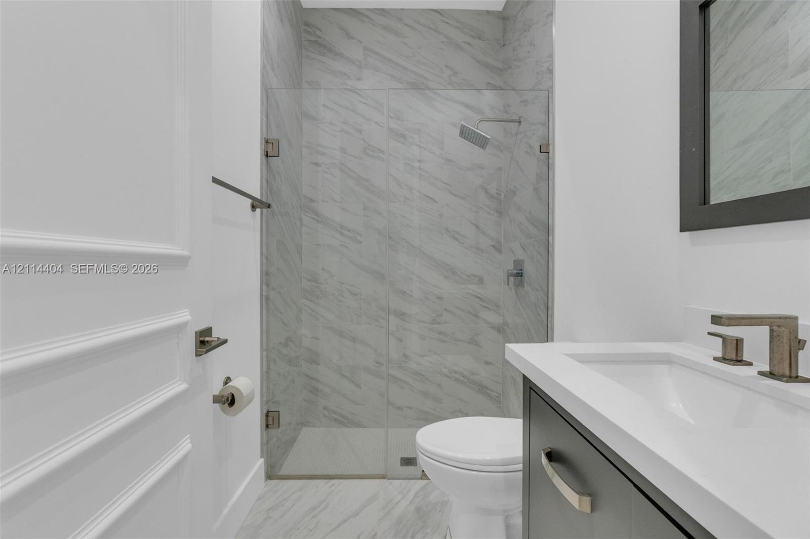 Bathroom featuring a walk-in shower with stone-patterned tile, frameless glass enclosure, wall-mounted rainfall showerhead, and a vanity with a white countertop and dark cabinetry