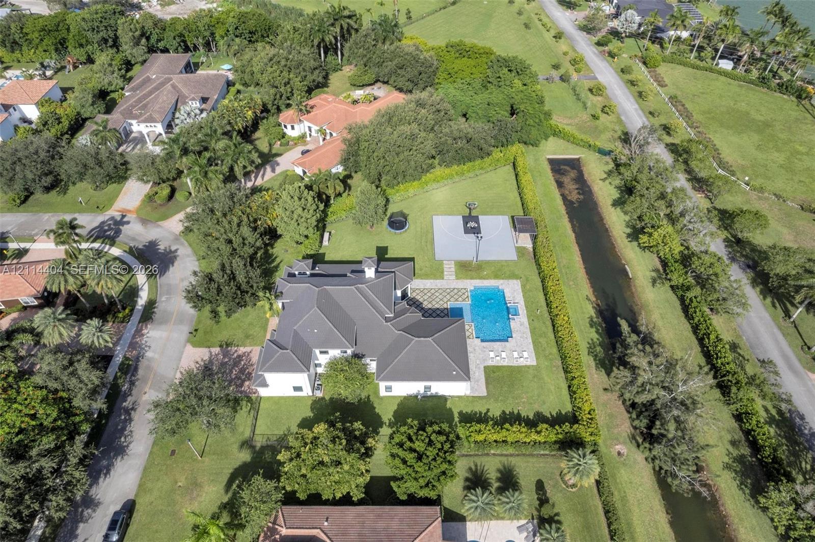 Aerial perspective featuring a multi-gabled roofline, a private swimming pool with patterned stone decking, a paved sports court, and mature landscaping