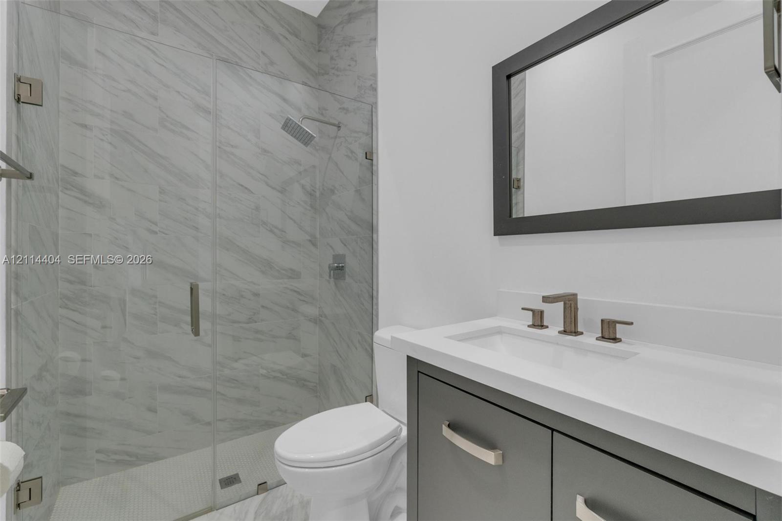 Modern vanity with a rectangular sink, stone-finish countertop, and dark-toned cabinetry, paired with a glass-enclosed shower featuring large-format tile surrounds, a rain-style showerhead, and a framed mirror