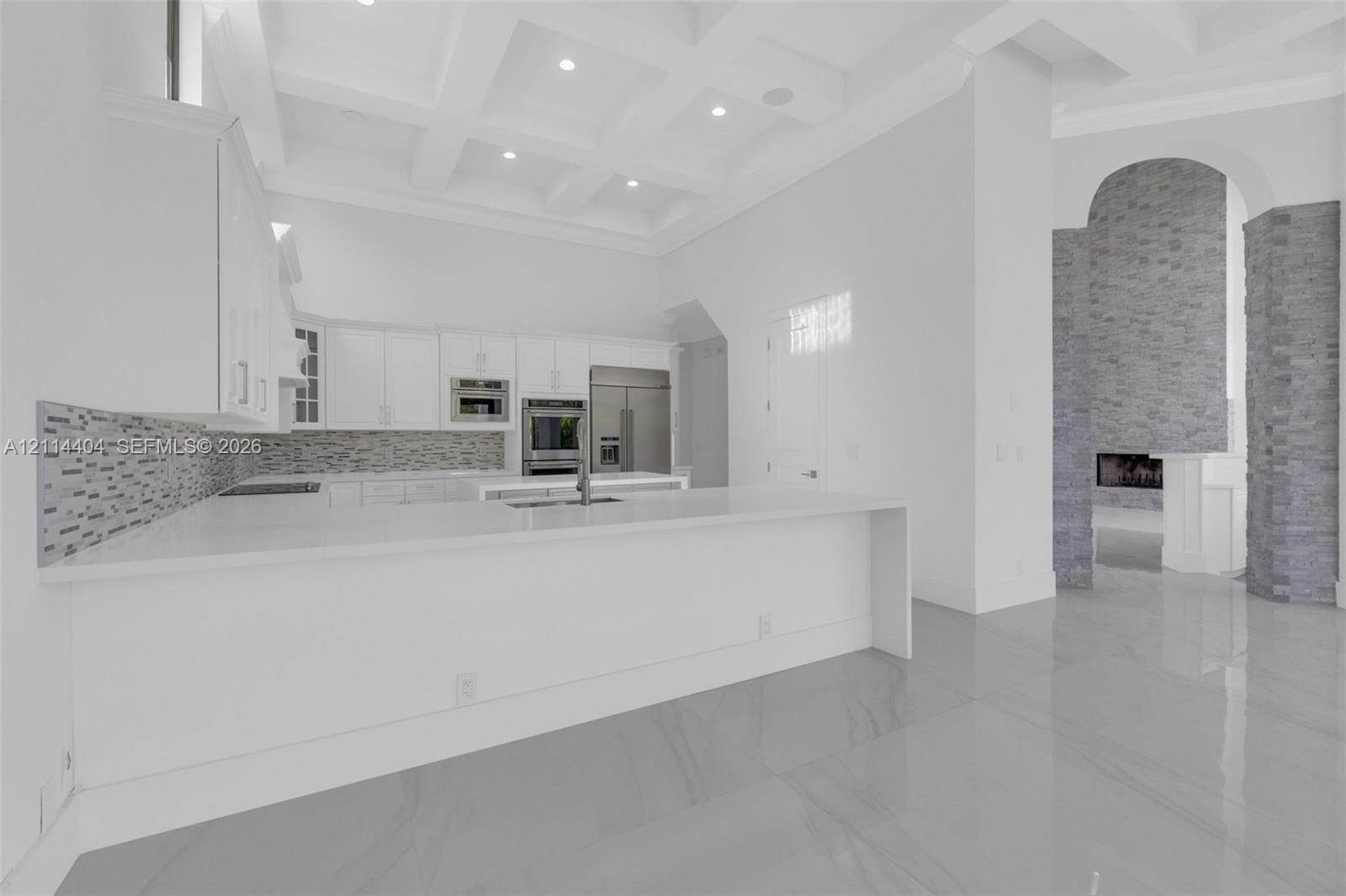Kitchen featuring custom cabinetry, a stone-tile backsplash, expansive solid-surface island, and coffered ceilings, complemented by a floor-to-ceiling stone fireplace surround