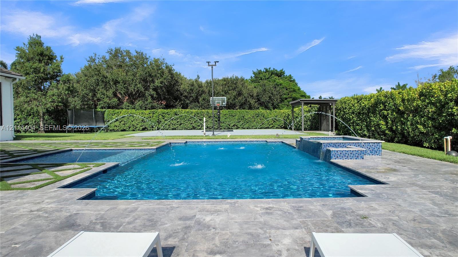 Backyard featuring an in-ground pool with water features, an attached spillover spa, stone-finish paver patio, and a concrete-surfaced sports court
