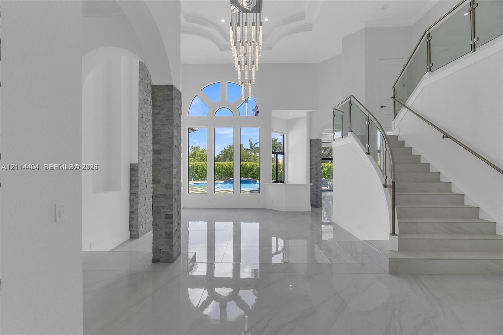 Grand foyer featuring polished stone flooring, a multi-tiered crystal chandelier, vaulted ceilings with recessed molding, a curved staircase with glass railings, and floor-to-ceiling arched windows with stacked stone pillars Grand foyer featuring polished stone flooring, a multi-tiered crystal chandelier, vaulted ceilings with recessed molding, a curved staircase with glass railings, and floor-to-ceiling arched windows with stacked stone pillars