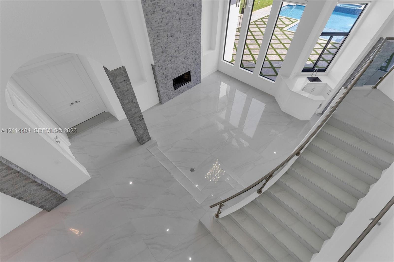 High-ceiling interior featuring oversized polished tile flooring, full-height stone fireplace surround, curved staircase with metal railing, and floor-to-ceiling windows