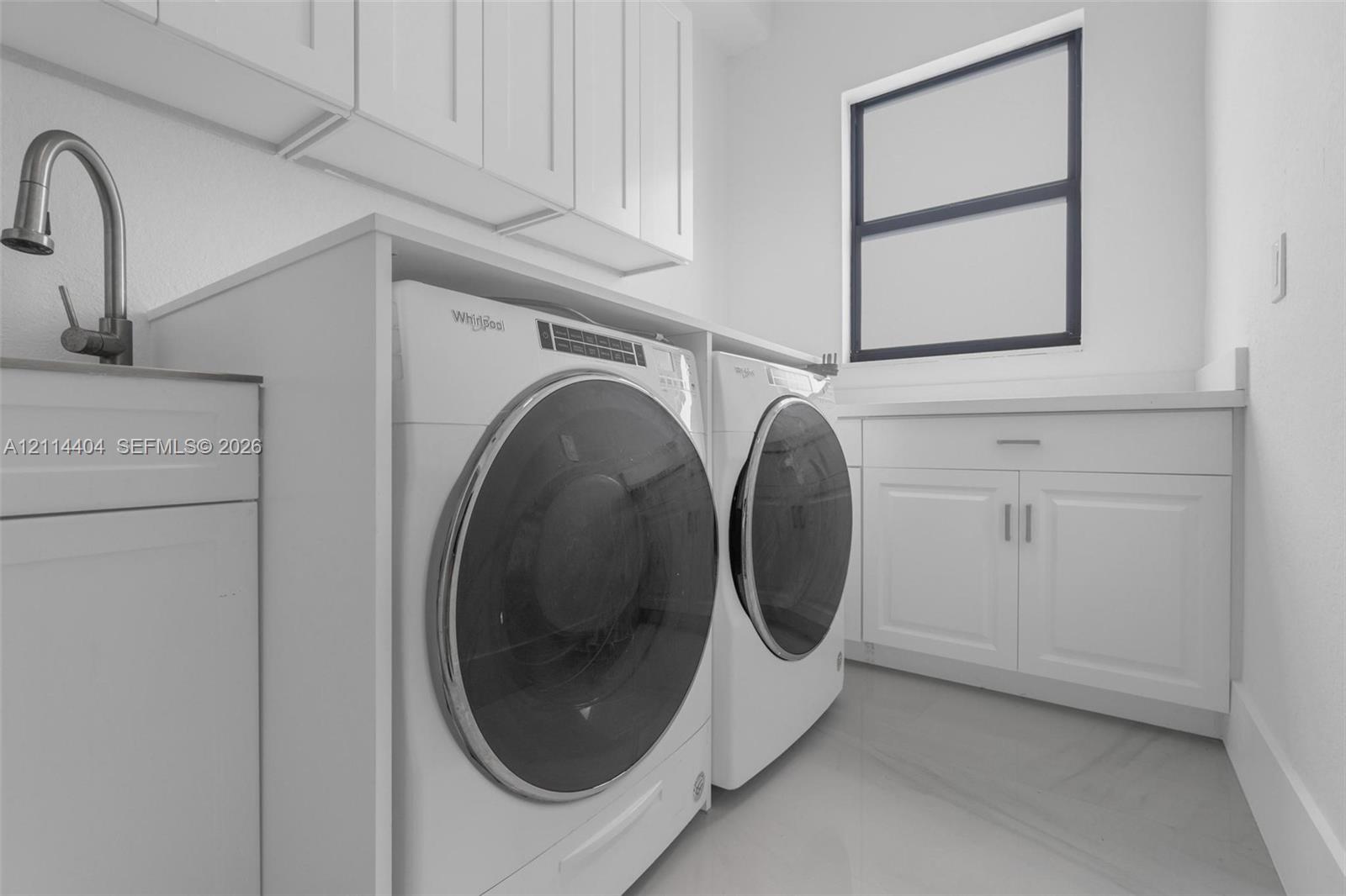 Laundry area featuring white shaker-style cabinetry, a built-in sink with a high-arch faucet, solid-surface countertops, and large-format tile flooring