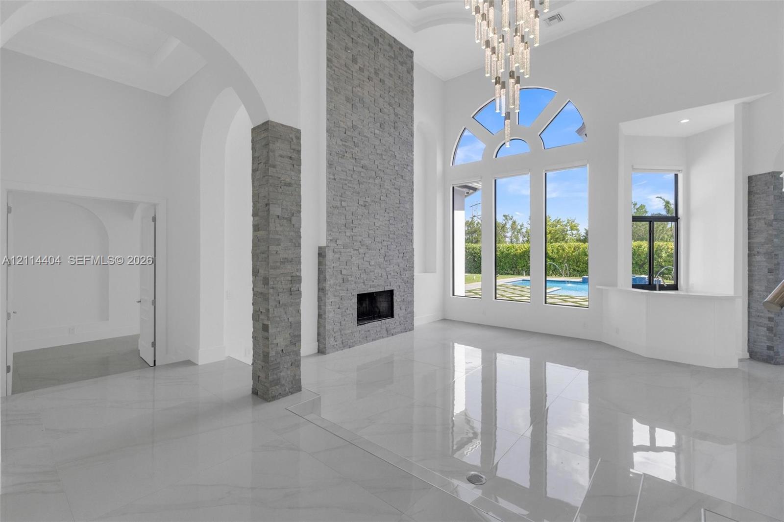 Double-height ceilings featuring an expansive stacked-stone fireplace, polished tile flooring, arched architectural detailing, a multi-tiered crystal chandelier, and large-scale windows with exterior pool exposure Double-height ceilings featuring an expansive stacked-stone fireplace, polished tile flooring, arched architectural detailing, a multi-tiered crystal chandelier, and large-scale windows with exterior pool exposure