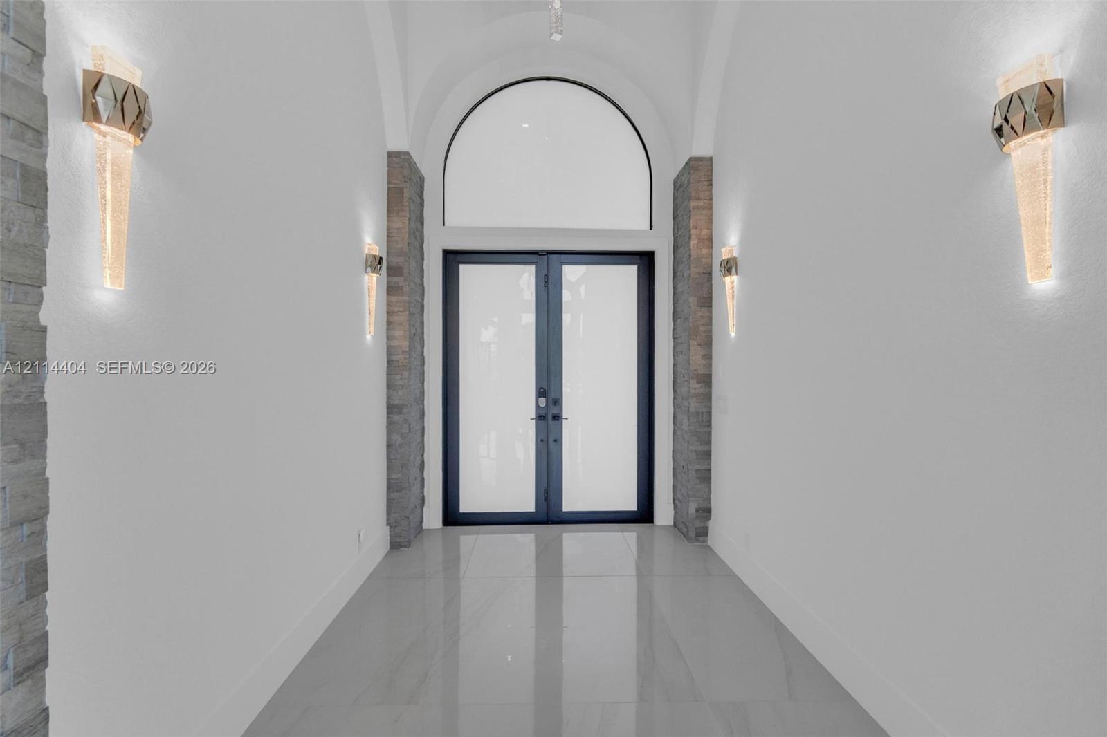 Entryway featuring arched architectural detailing, double glass-panel doors, stone-finish columns, and large-format tile flooring