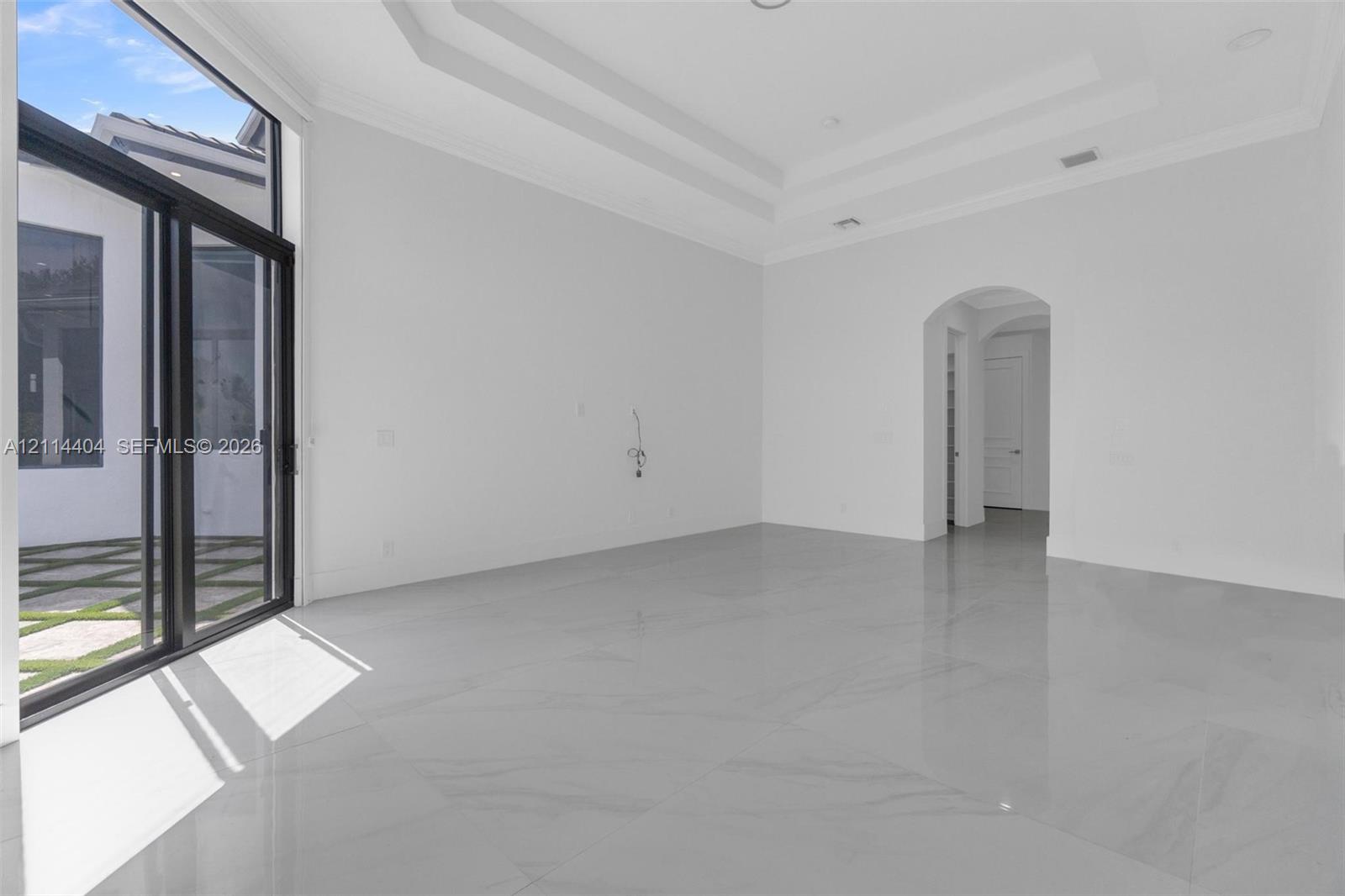 High-ceiling interior featuring a multi-tiered tray ceiling, floor-to-ceiling glass sliding doors, polished stone-finish flooring, and an arched entryway