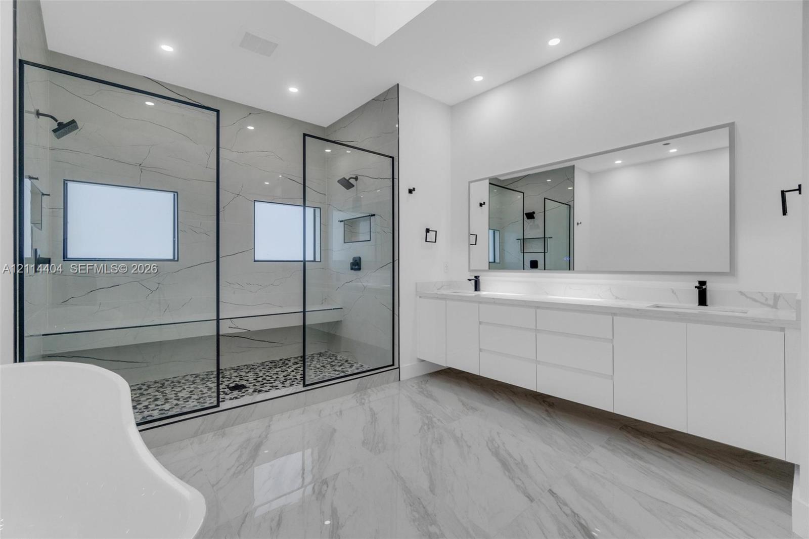 Double vanity with integrated sinks, expansive walk-in shower featuring a stone-tile floor and matte black hardware, large frameless mirror, and recessed ceiling lighting
