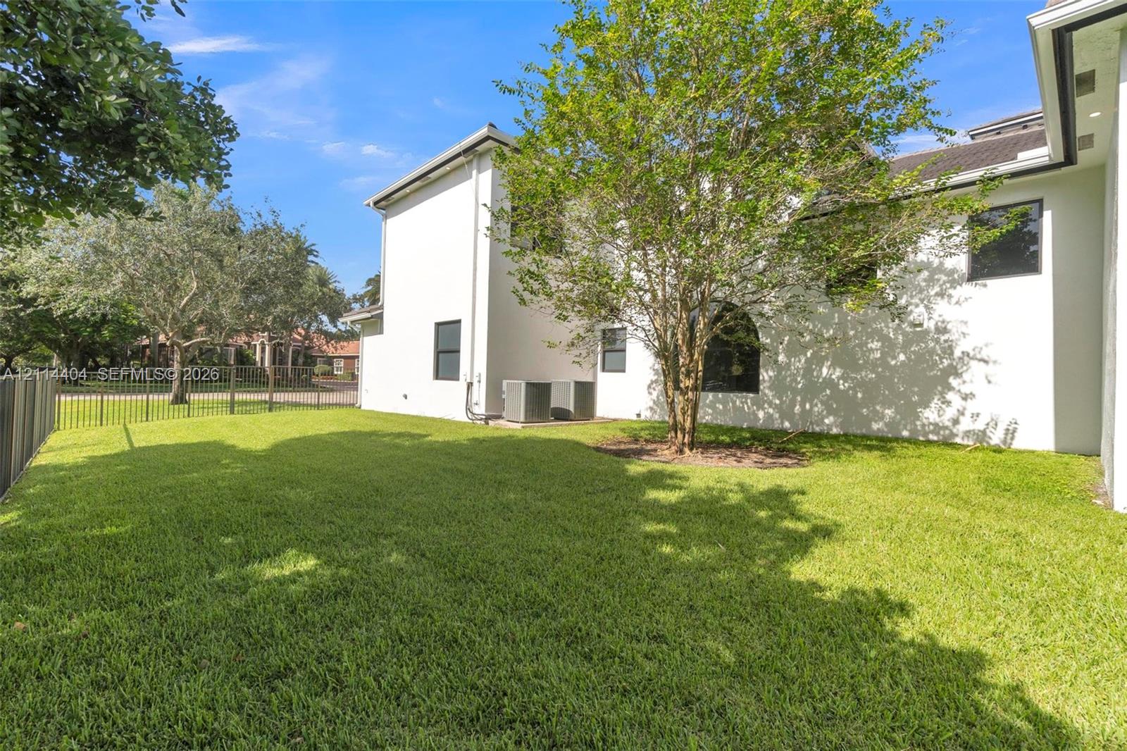 Exterior side yard featuring a manicured lawn, metal perimeter fencing, light-toned smooth exterior wall finish, and mature shade trees