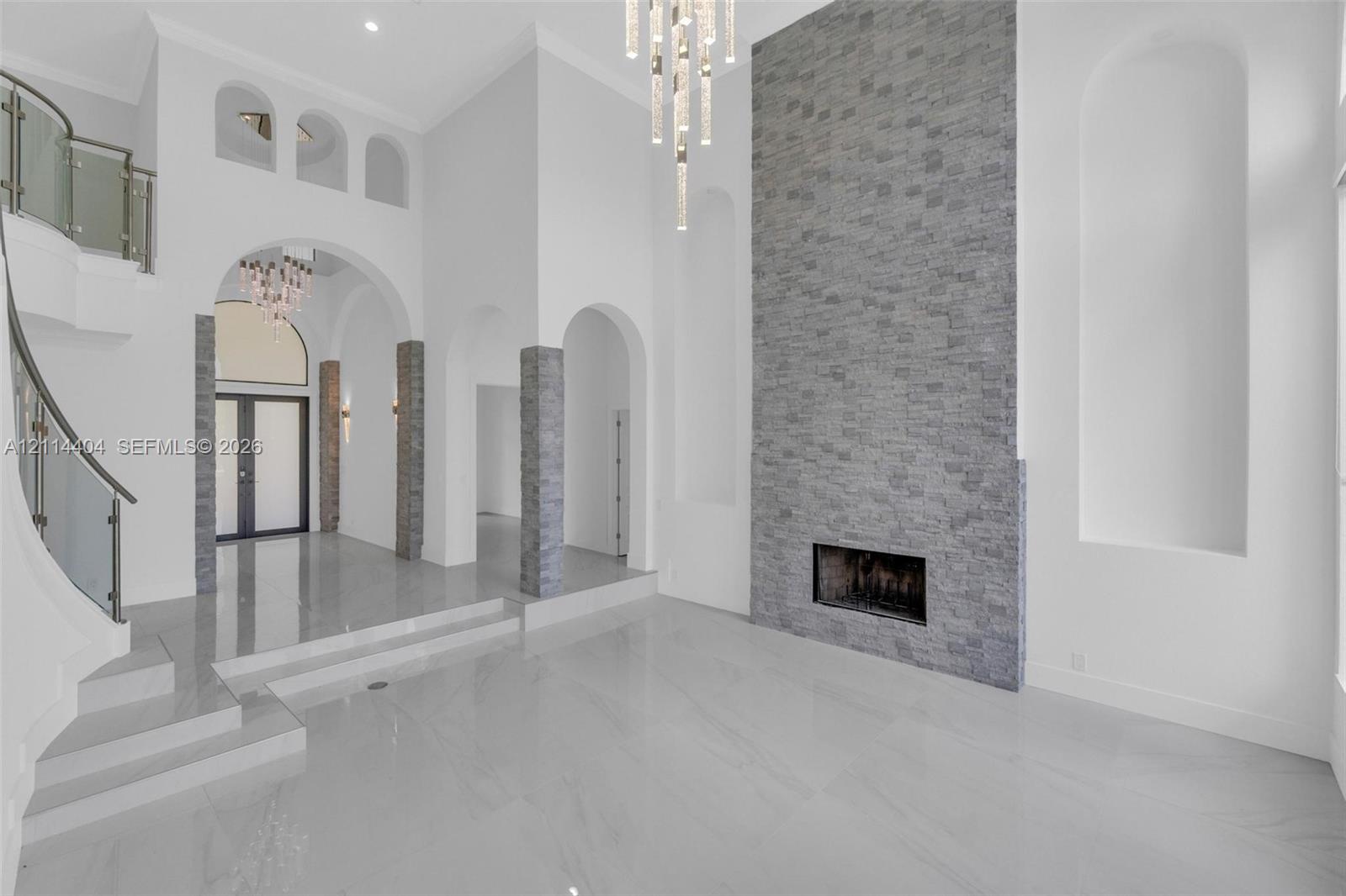 High-ceiling foyer with polished tile flooring, stone-veneer fireplace wall, architectural archways, built-in display niches, and a curved staircase featuring a metal and glass railing High-ceiling foyer with polished tile flooring, stone-veneer fireplace wall, architectural archways, built-in display niches, and a curved staircase featuring a metal and glass railing