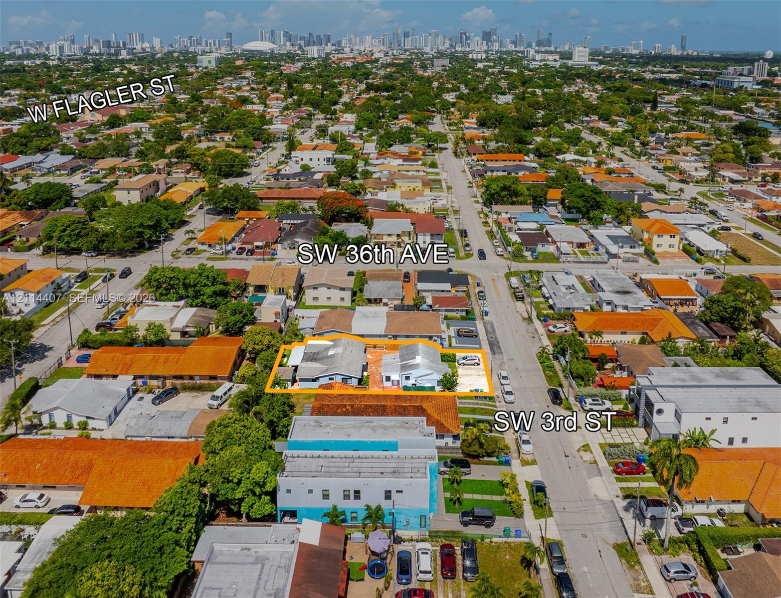 Aerial perspective of a corner lot situated at the intersection of SW 36th Avenue and SW 3rd Street, featuring two structures with light-colored roofs, paved parking areas, and proximity to the city skyline