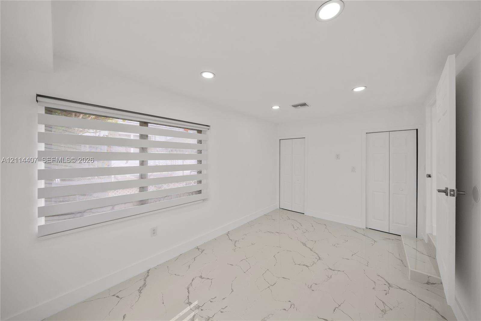 Bright room featuring large-format veined tile flooring, recessed ceiling lighting, dual closets with bi-fold doors, and a window with adjustable layered roller shades
