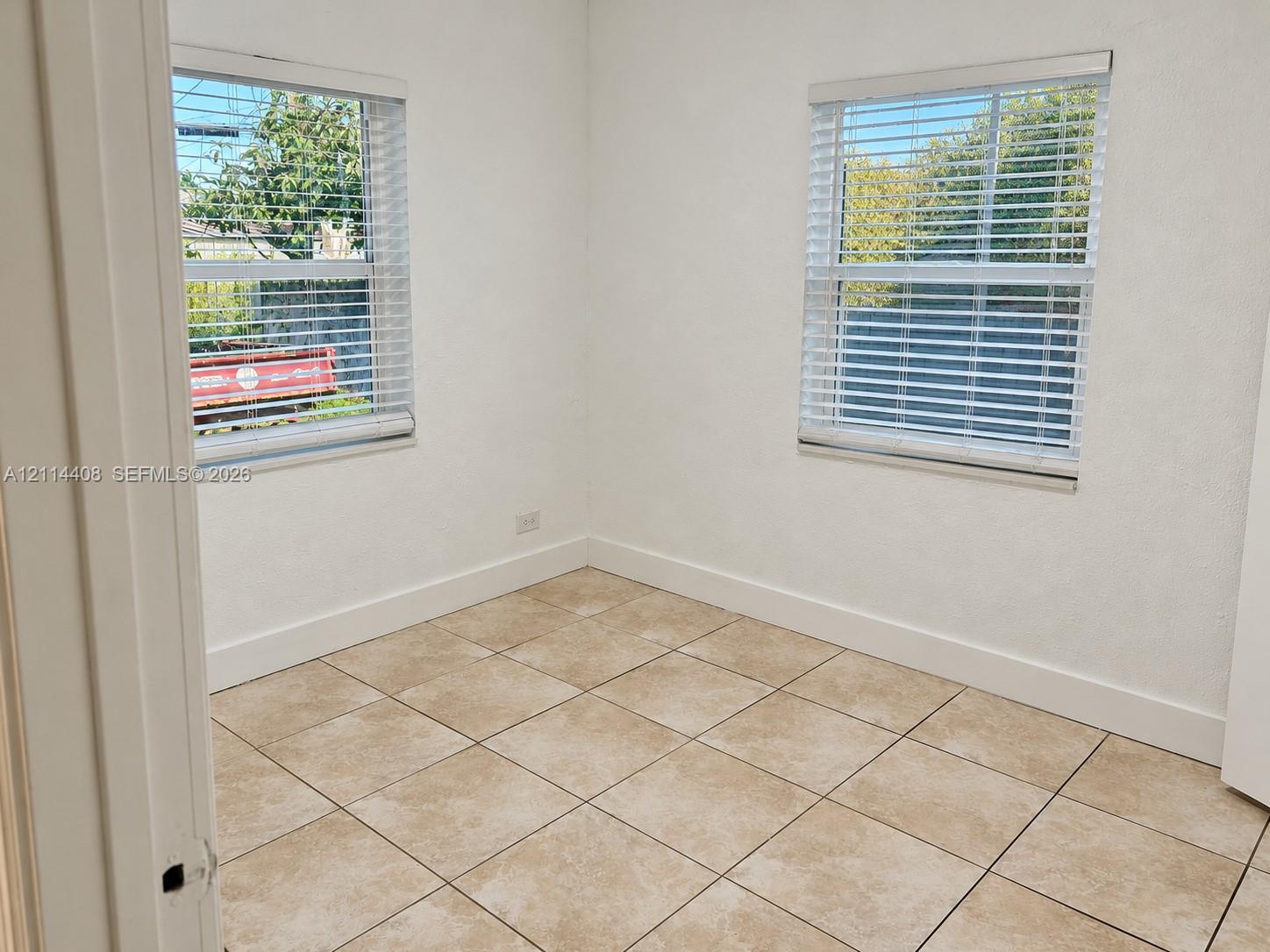 Room featuring neutral-toned tile flooring, two windows with horizontal blinds, and smooth-finish white walls