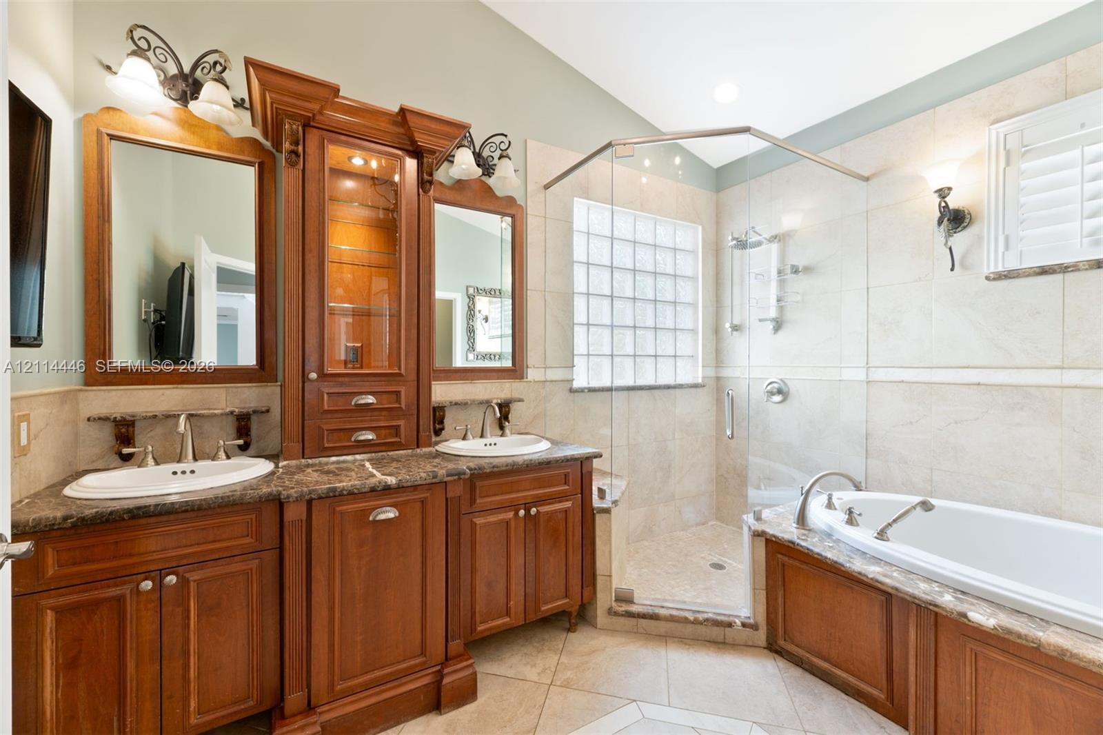 Bathroom featuring dual undermount sinks with stone countertops, cherry-finish cabinetry, a glass-enclosed shower with glass-block window, and a built-in soaking tub