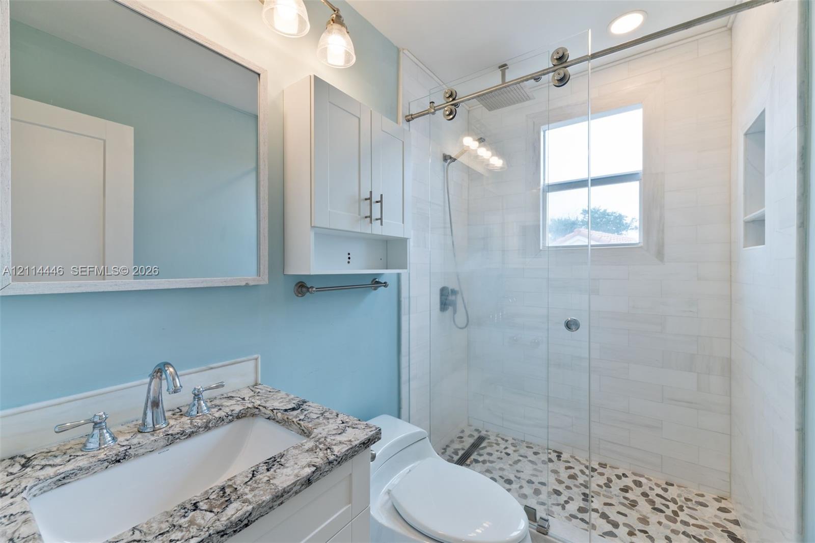 Bathroom featuring a stone-top vanity with an undermount sink, walk-in shower with a sliding glass door, pebble tile shower floor, and built-in recessed wall shelving