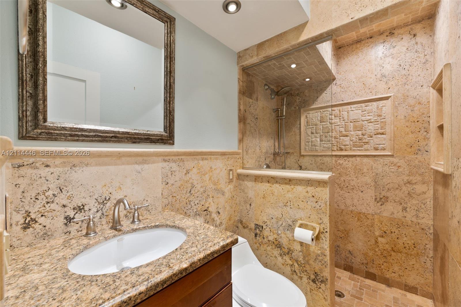 Bathroom featuring stone tile wainscoting and shower surround, stone countertop with an undermount sink, built-in shower niche, glass shower partition, and recessed ceiling lighting