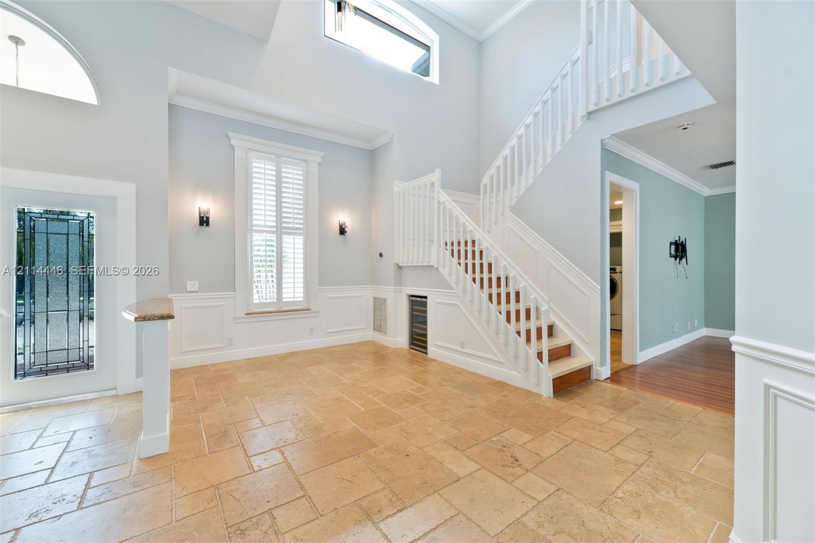 Grand foyer featuring vaulted ceilings, natural stone tile flooring, a staircase with custom white millwork, decorative wall paneling, and an integrated wine chiller