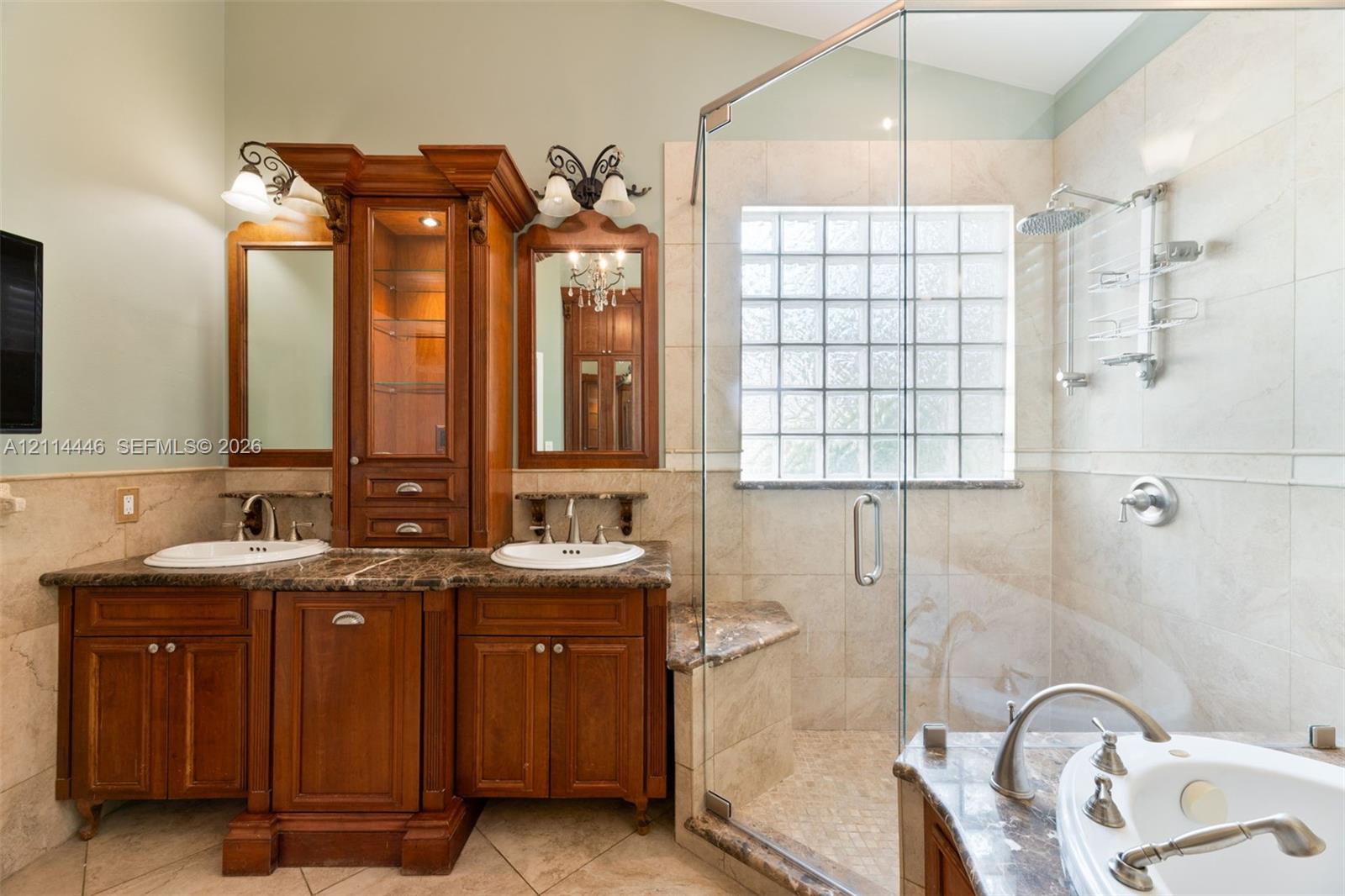 Dual-sink vanity with wood cabinetry, stone countertops, glass-enclosed shower featuring a built-in bench, glass block window, and soaking tub with deck-mounted faucet