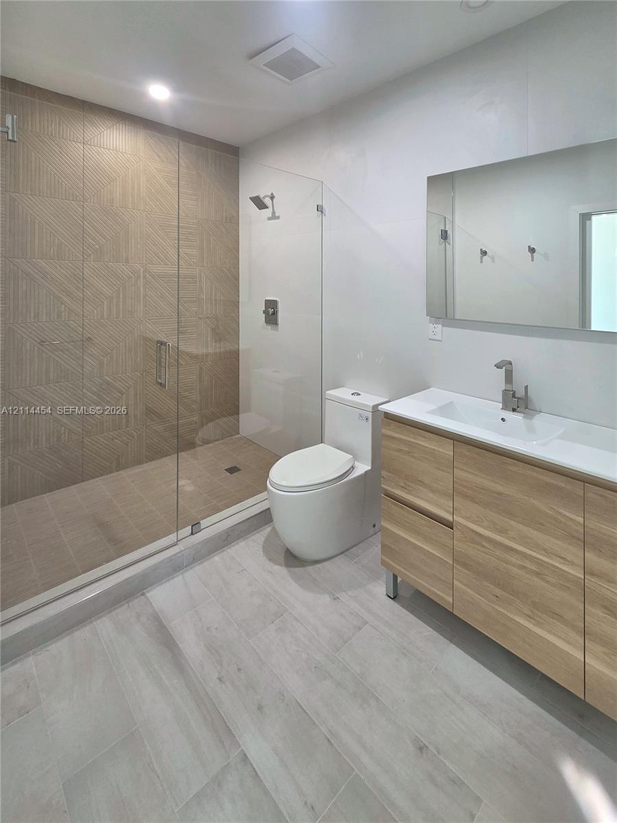 Walk-in shower with textured tile surround, frameless glass enclosure, wood-finish vanity, integrated rectangular sink, and neutral-tone large-format floor tile