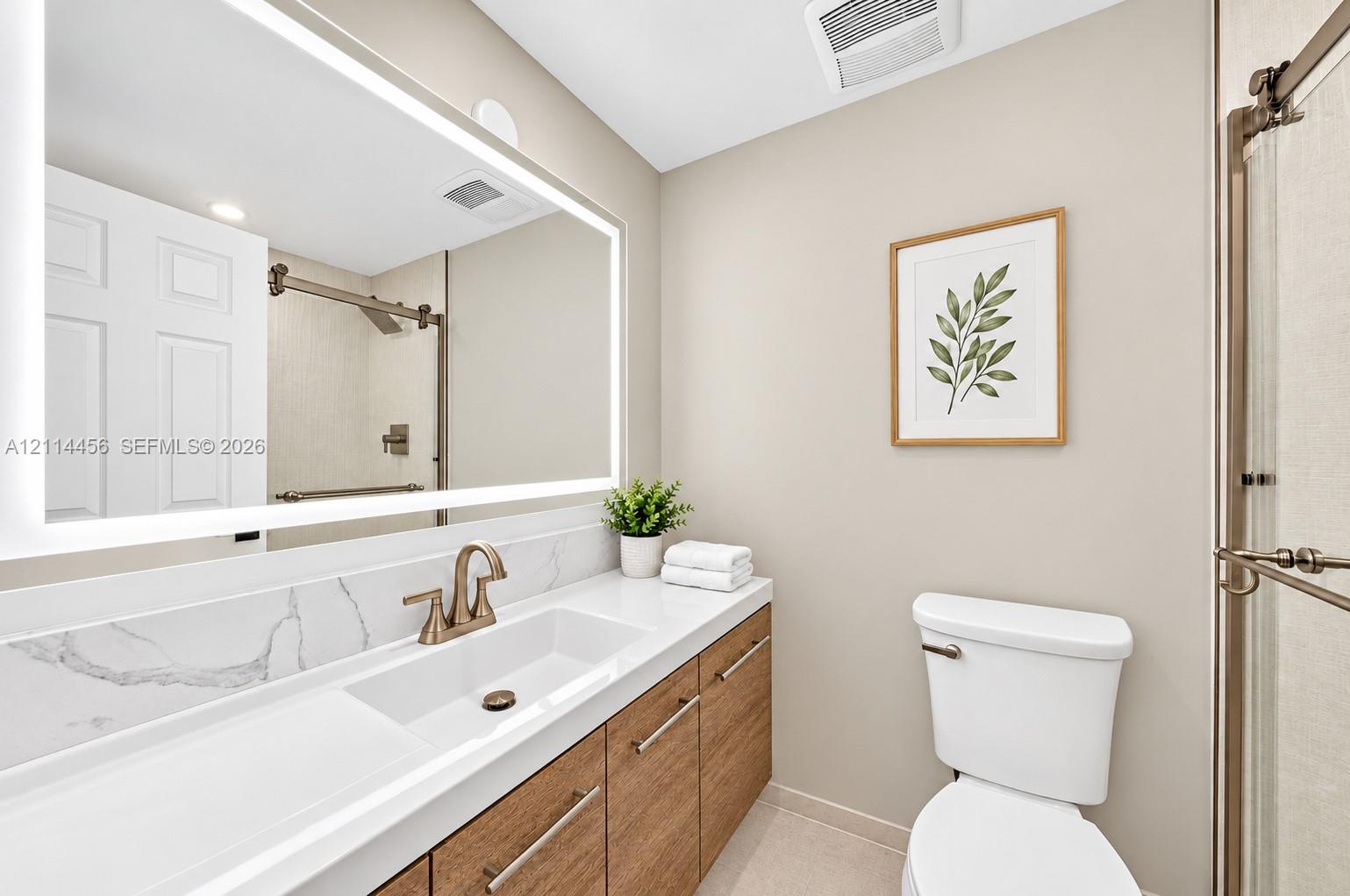 Bathroom featuring a wood-finish vanity with a stone-top integrated sink, LED-backlit mirror, brushed-finish faucet hardware, and a glass-enclosed shower