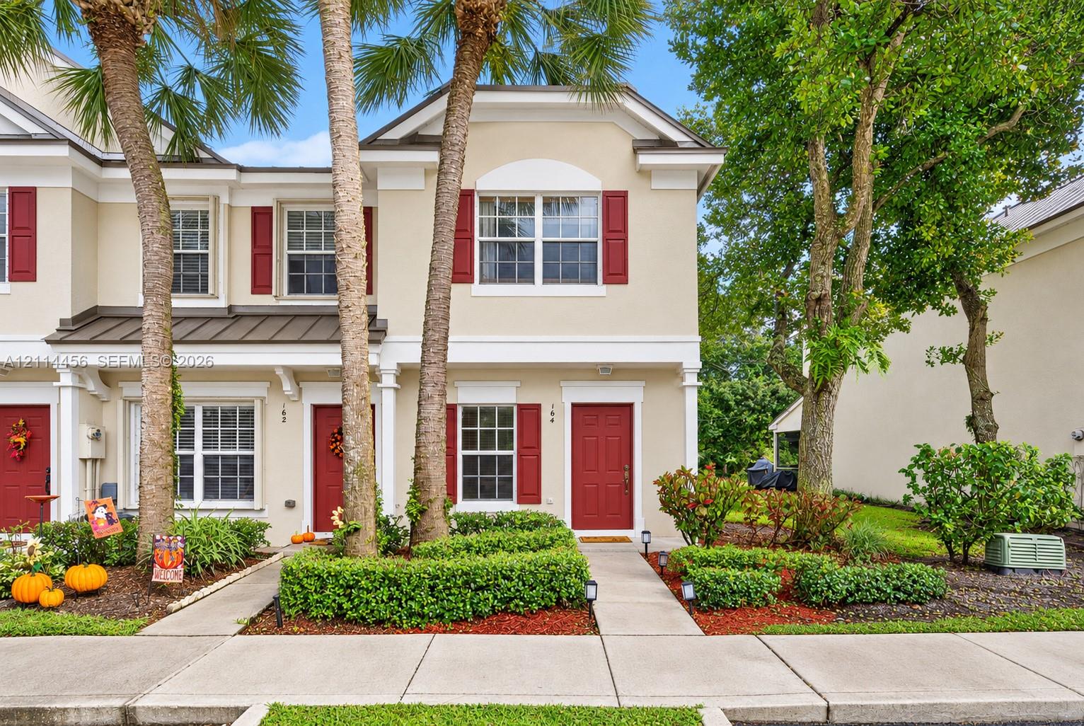 Two-story exterior featuring light-toned stucco, decorative shutters, a metal accent roof, and paved walkways surrounded by manicured landscaping and mature trees