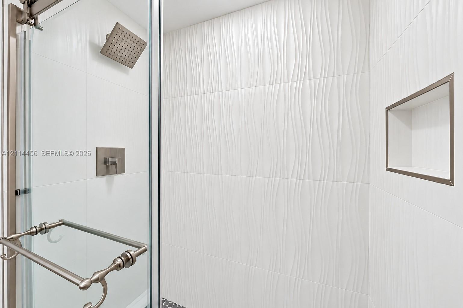 Walk-in shower featuring textured white wall tile, rain-style shower head, brushed metal fixtures, built-in recessed wall niche, and glass door enclosure