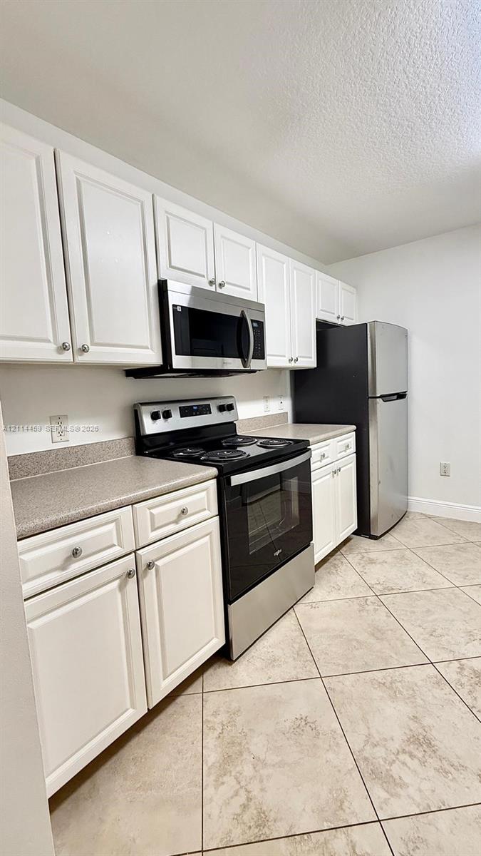 Kitchen featuring white shaker-style cabinetry, neutral solid-surface countertops, tile flooring, and stainless steel appliances including a built-in microwave, range, and refrigerator