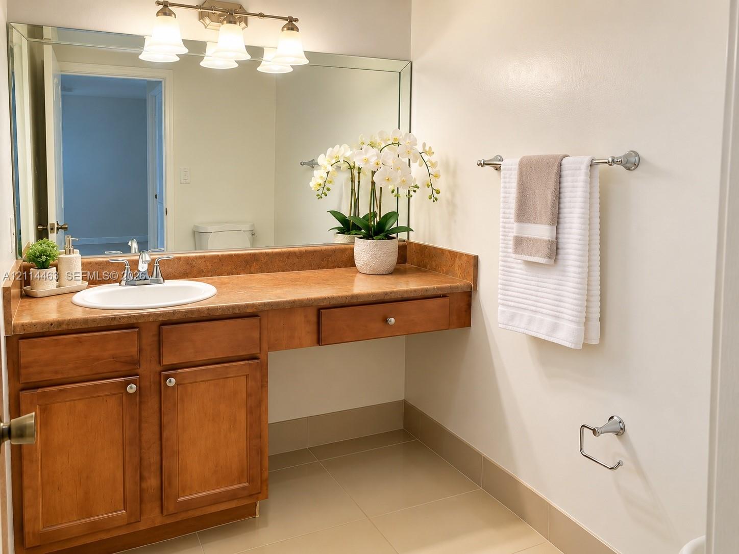 Bathroom vanity featuring warm-toned cabinetry, an integrated stone-finish countertop with a circular drop-in sink, a large wall-mounted vanity mirror, a three-light fixture, and neutral tile flooring Bathroom vanity featuring warm-toned cabinetry, an integrated stone-finish countertop with a circular drop-in sink, a large wall-mounted vanity mirror, a three-light fixture, and neutral tile flooring