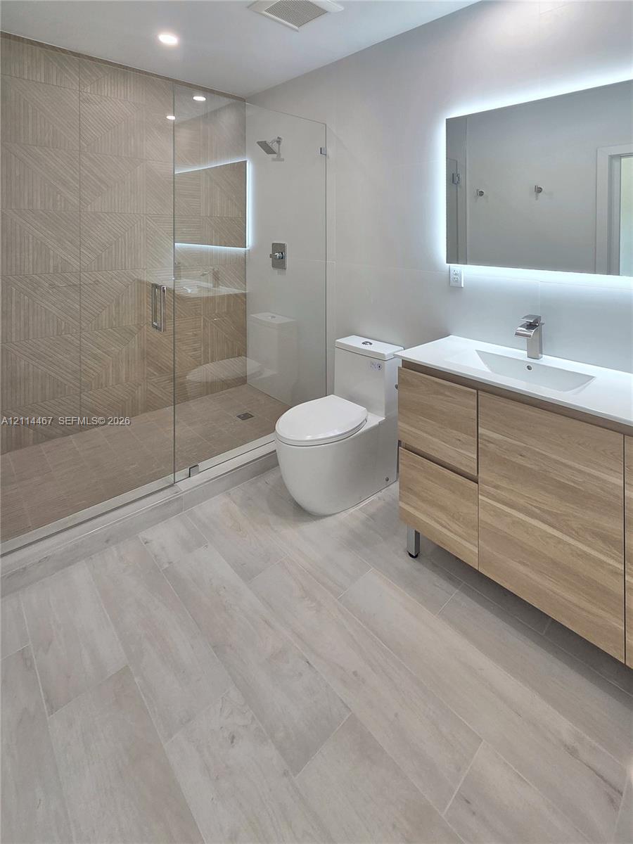 Modern bathroom featuring a wood-finish floating vanity, integrated rectangular sink, frameless glass shower enclosure with textured tile wall, and backlit mirror
